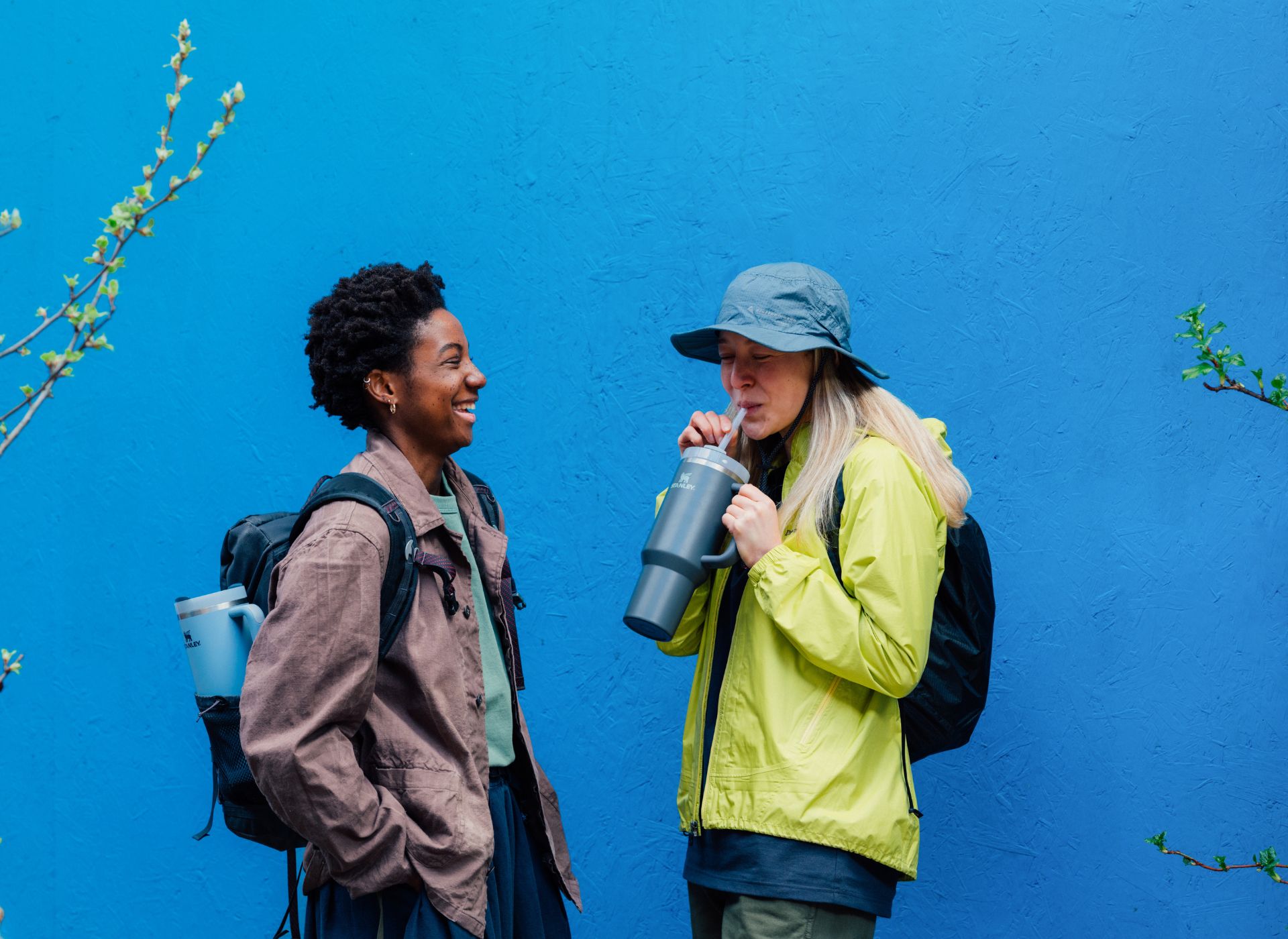 Two women stood laughing in front of blue wall. One is holding a Stanley quencher