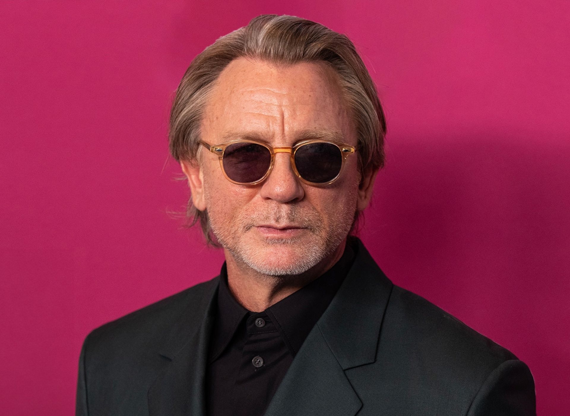 Daniel Craig wearing sunglasses pictured in front of pink backdrop